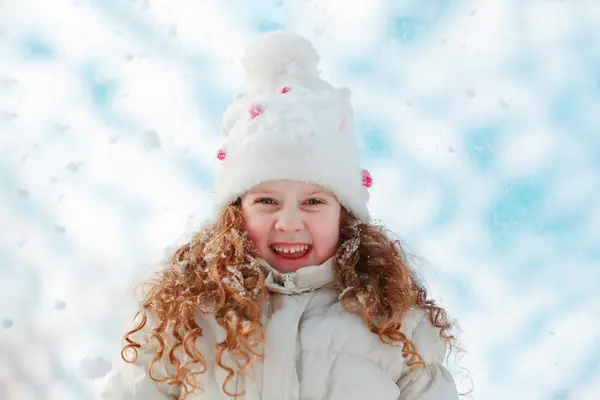 Jente med brunt krøllet hår nedenfor en hvit lue og med hvit jakke og snø i bakgrunnen, som illustrasjonsbilde for beskrivelsen av Barnas Holmenkolldag på Østre Greverud gård
