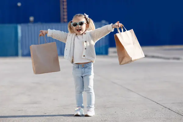 Liten jente med solbriller og fletter som strekker ut armene med en papirpose i hver hånd og et stort smil, som illustrasjonsbilde for beskrivelsen av barnas dag i Vestby kulturkvartal