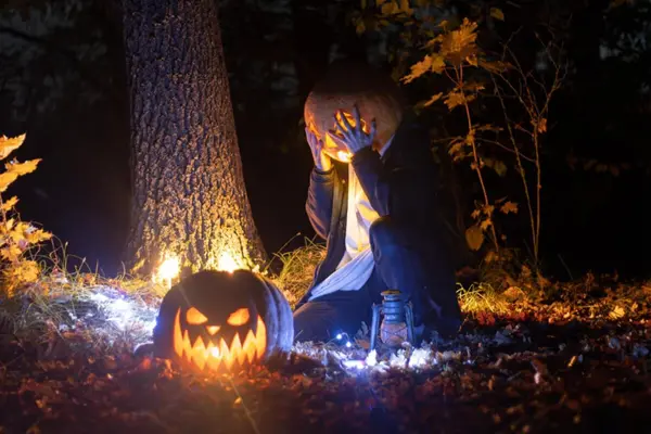 En person med gresskarhode sitter ved et tre i mørket omgitt av lysende gresskar og lykter under en Halloween-feiring i skogen