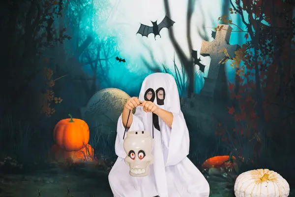 Et barn utkledd som spøkelse holder en hodeskallelykt foran et halloween-inspirert bakteppe med gresskar, flaggermus og gravstener