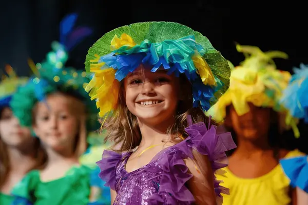 Jente med stor hatt full av fargede bånd og tyll og en fargerik kjole omgitt av andre barn i fargede lette drakter, som illustrasjon til beskrivelsen av barnelørdag i Venterommet på Oasen