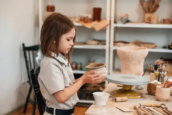 Et barn former en keramikkbolle i leire på et verkstedbord, omgitt av dreieskive, verktøy og ferdige keramikkarbeider, som illustrasjonsbilde for beskrivelsen av keramikkverksted på Deichman Romsås i vinterferien