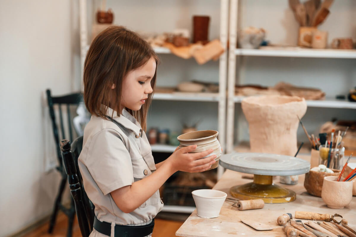 Et barn former en keramikkbolle i leire på et verkstedbord, omgitt av dreieskive, verktøy og ferdige keramikkarbeider, som illustrasjonsbilde for beskrivelsen av keramikkverksted på Deichman Romsås i vinterferien