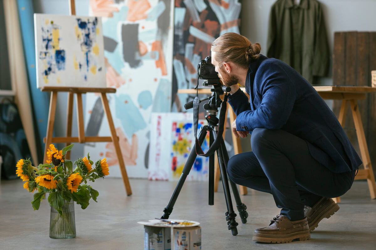 En mann fotograferer et stilleben med solsikker i et kunstatelier omgitt av malerier og staffelier, som illustrasjonsbilde for beskrivelsen av Oslo Open
