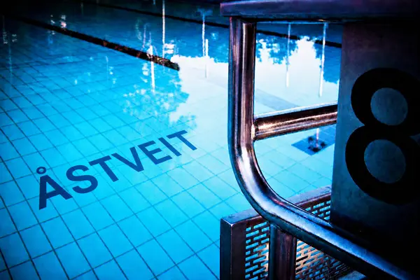Innendørs svømmebasseng, bilde av deler av vannspeilet og en startkloss med nummer åtte på siden og ÅSTVEIT skrevet med store bokstaver på bunnen, som illustrasjonsbilde for beskrivelsen av Åstveit svømmehall