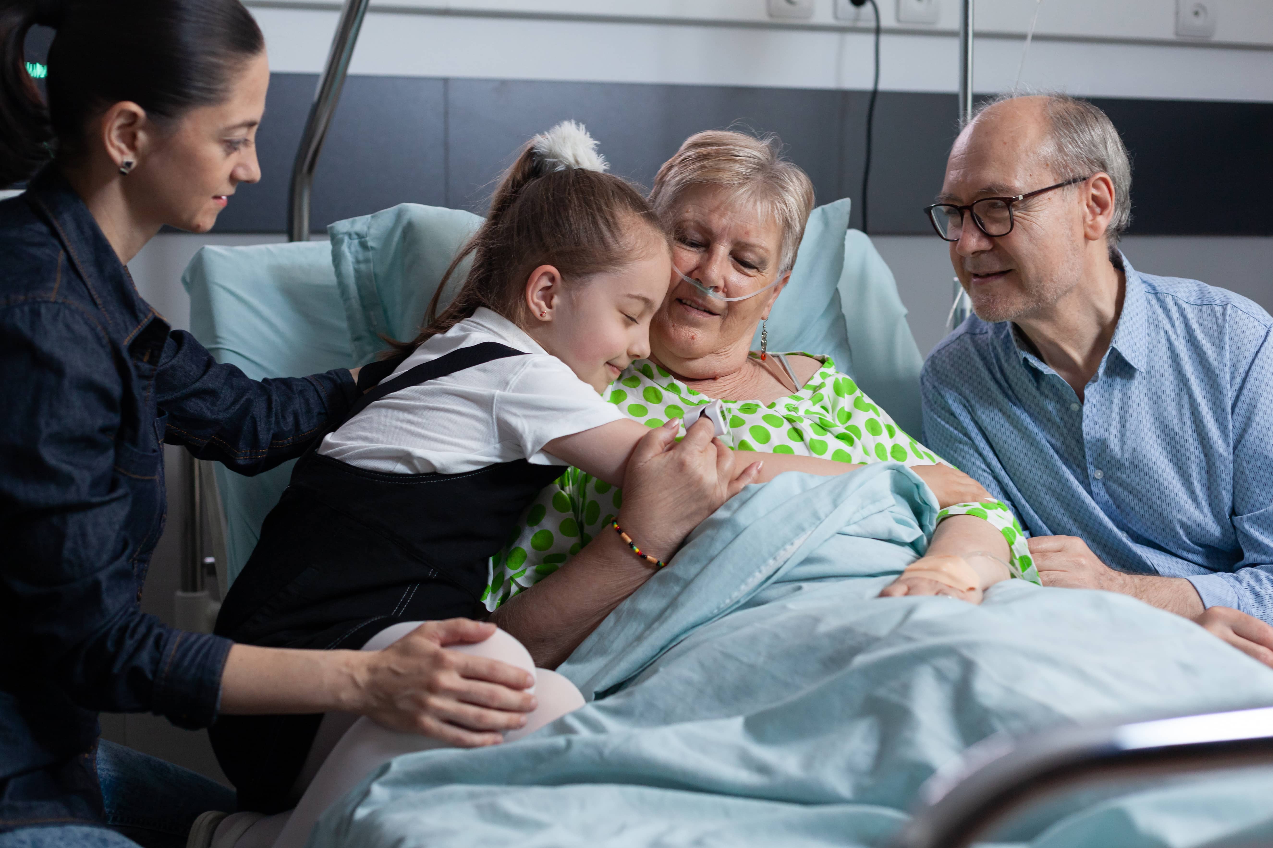 Elderly woman in hospital bed with oxygen tube, embracing a young girl while a woman and man look on supportively.