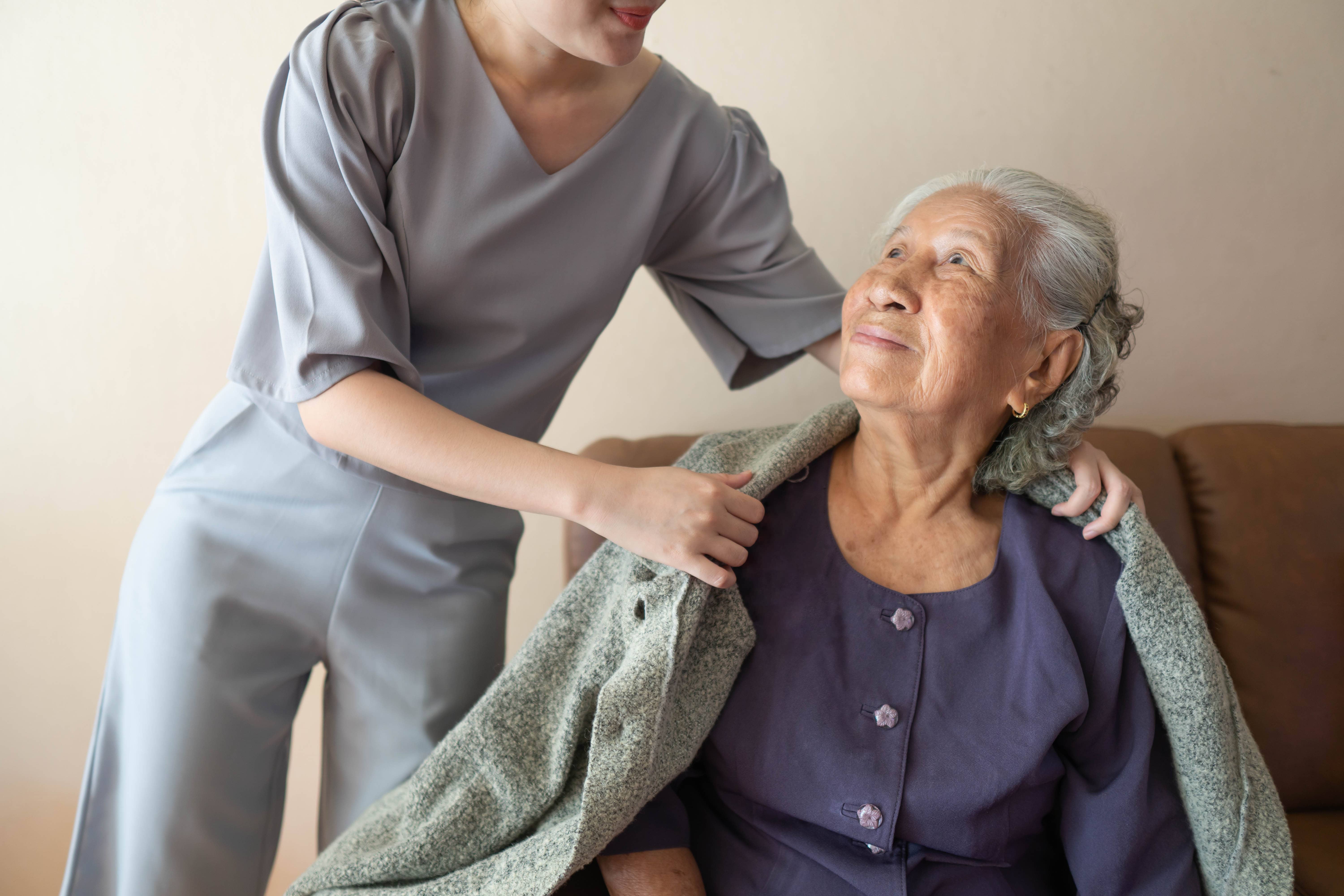 Younger person helping an elderly woman put on a gray coat indoors.