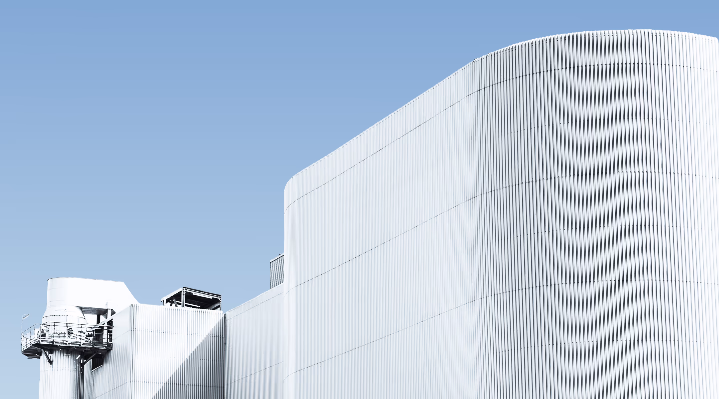Large white industrial building with a curved corrugated metal exterior under a clear blue sky.