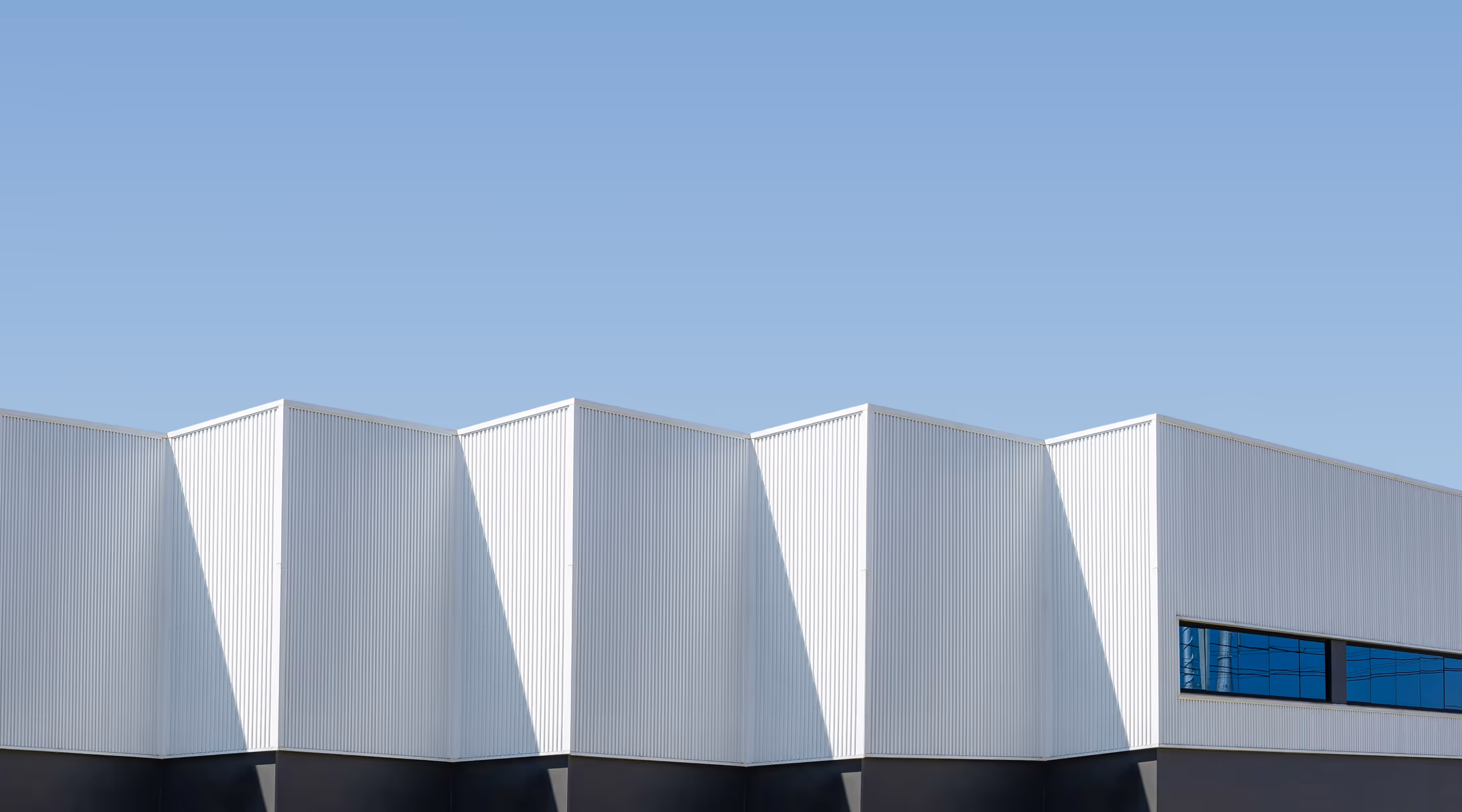 Modern white industrial building with angular roof sections under clear blue sky.