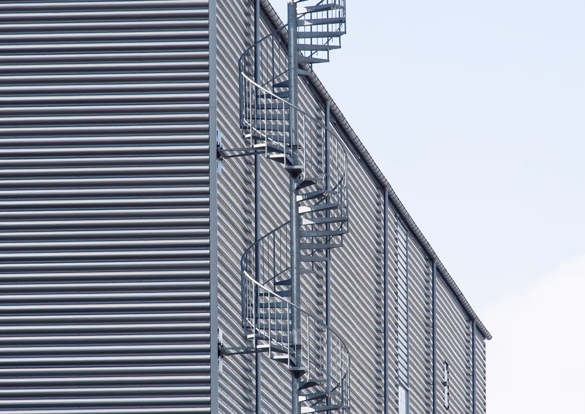Metal spiral staircase attached to the corrugated metal wall of an industrial building against a clear sky.