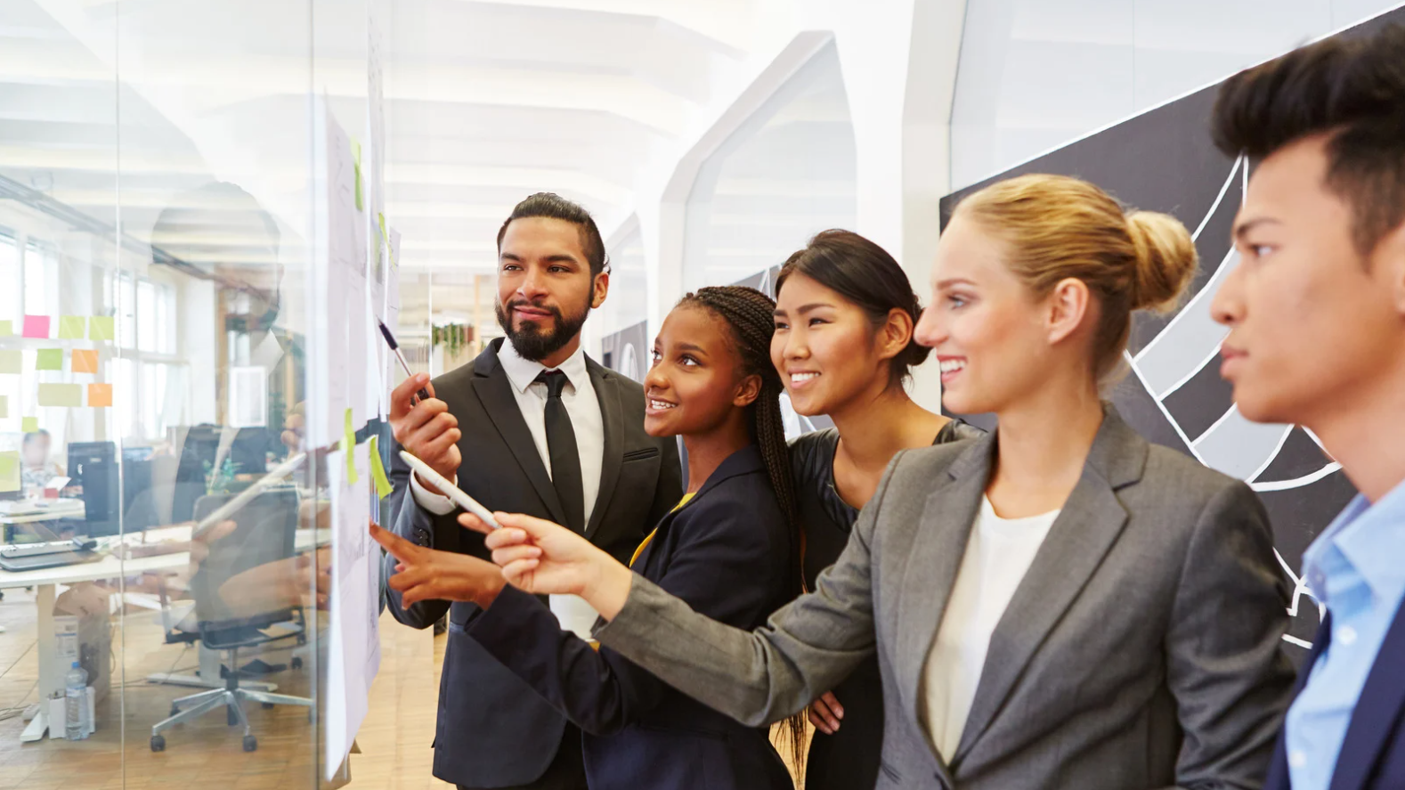 A diverse group of five business professionals collaborating and discussing ideas on sticky notes on a glass wall in a modern office.