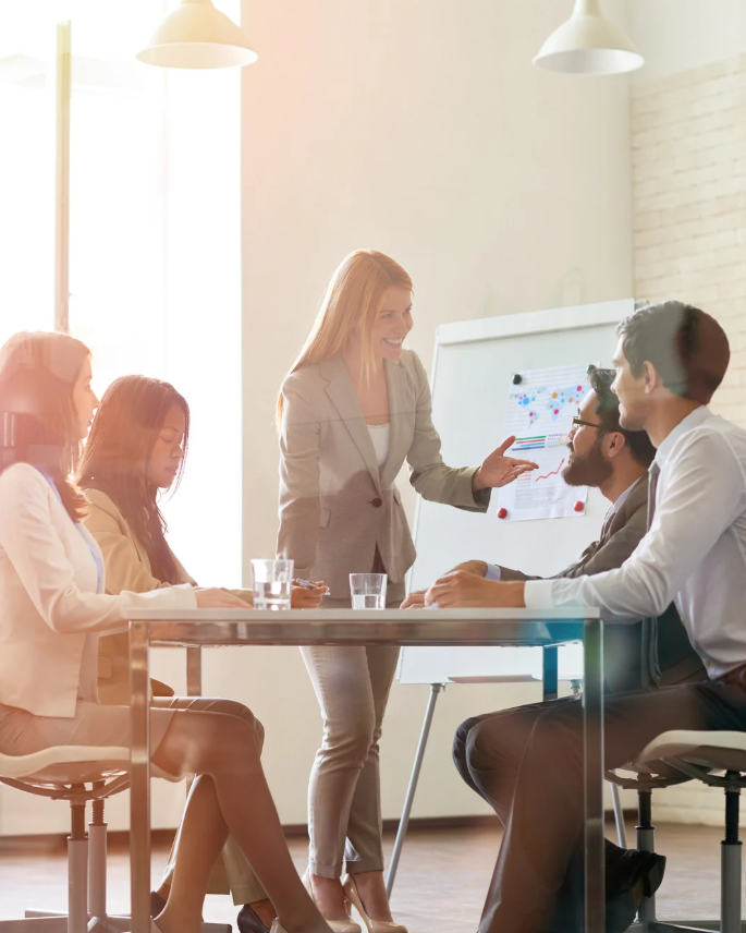 Businesswoman presenting data on a flip chart to three seated colleagues in a bright office meeting room.