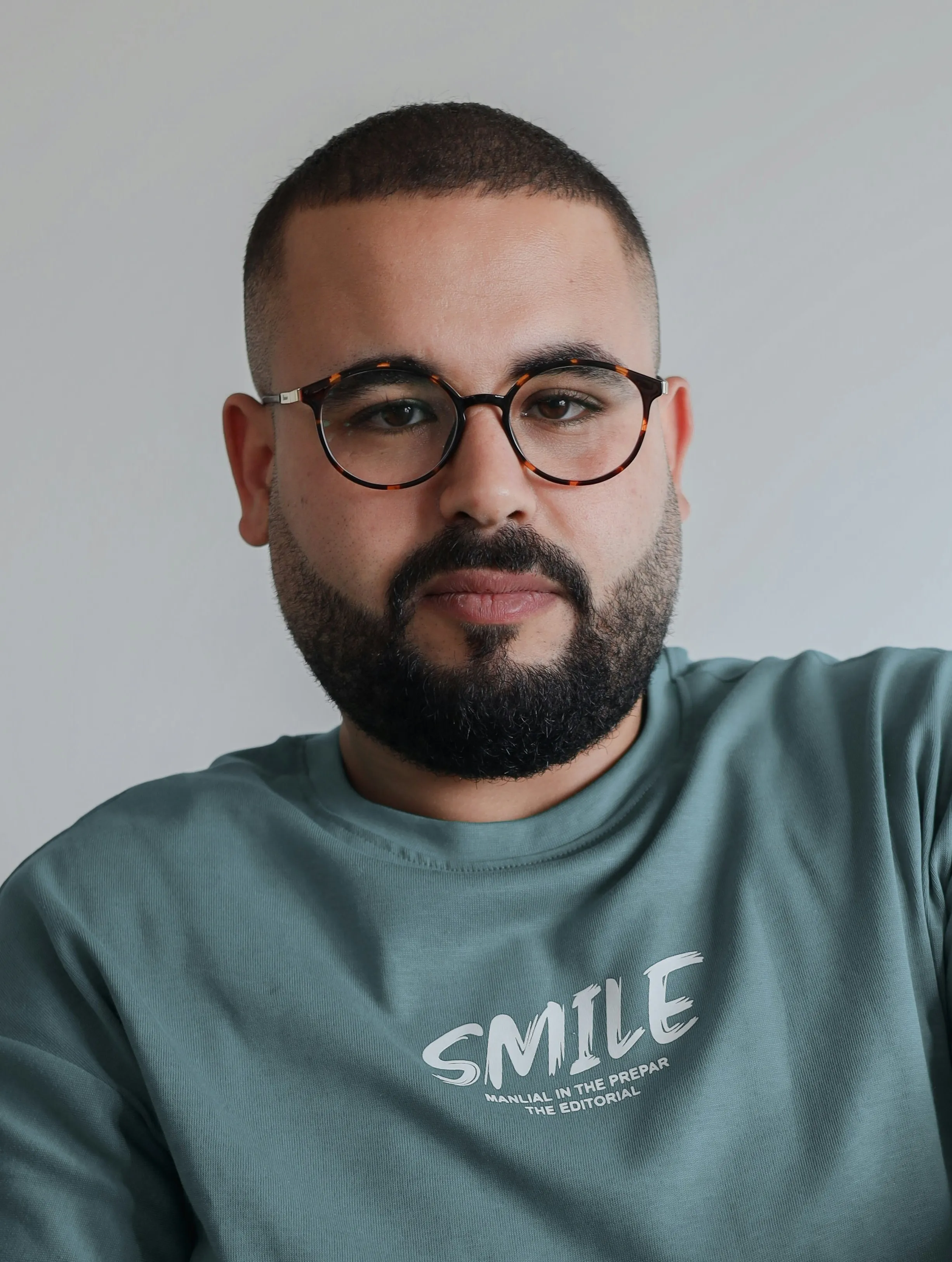 Bearded man wearing round tortoiseshell glasses and a teal shirt with the word 'SMILE' printed on it, against a plain light background.