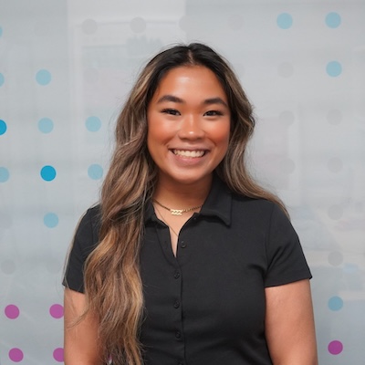 Smiling woman with long wavy hair wearing a black button-up shirt standing against a light background with blue and purple dots.