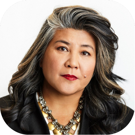 Portrait of a woman with long black and gray hair, red lipstick, wearing a black blazer and a statement necklace, against a white background.