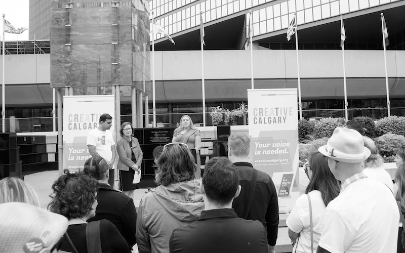 Group of people gathered outdoors listening to three speakers standing near Creative Calgary banners in an urban plaza.