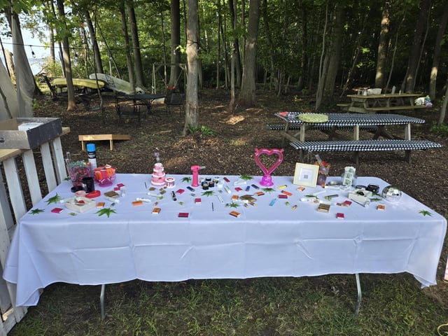 Outdoor table with a white tablecloth displaying various cannabis items in a messy setup, set in a wooded area with picnic tables in the background.
