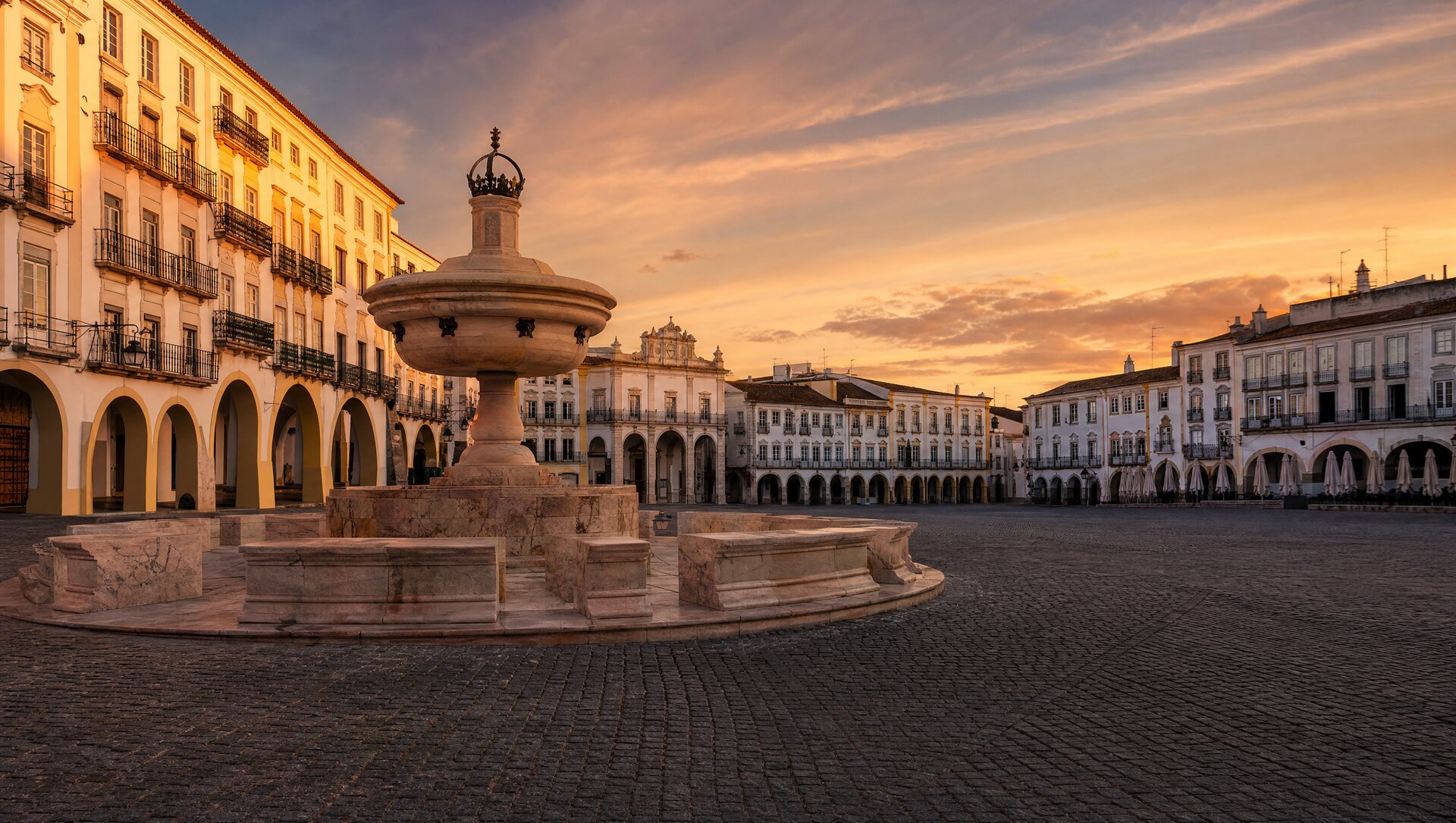 Praça do Giraldo à Évora au crépuscule, grande place pavée bordée d'arcades blanches, fontaine de marbre au centre, lumière dorée de fin de journée sur les façades chaulées de l'Alentejo
