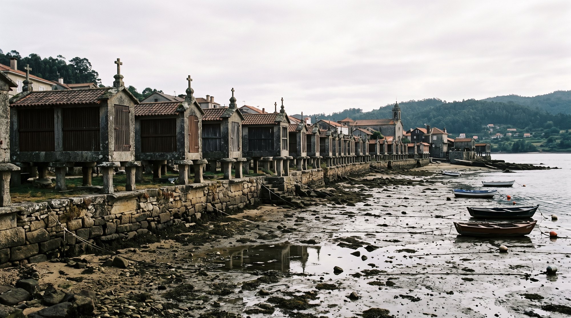 Alignement d'une trentaine d'hórreos de granit de Combarro dressés sur pieds de pierre au bord de la ría de Pontevedra, portes tournées vers la mer, petites croix au sommet, lumière grise et argentée de Galice, marée basse