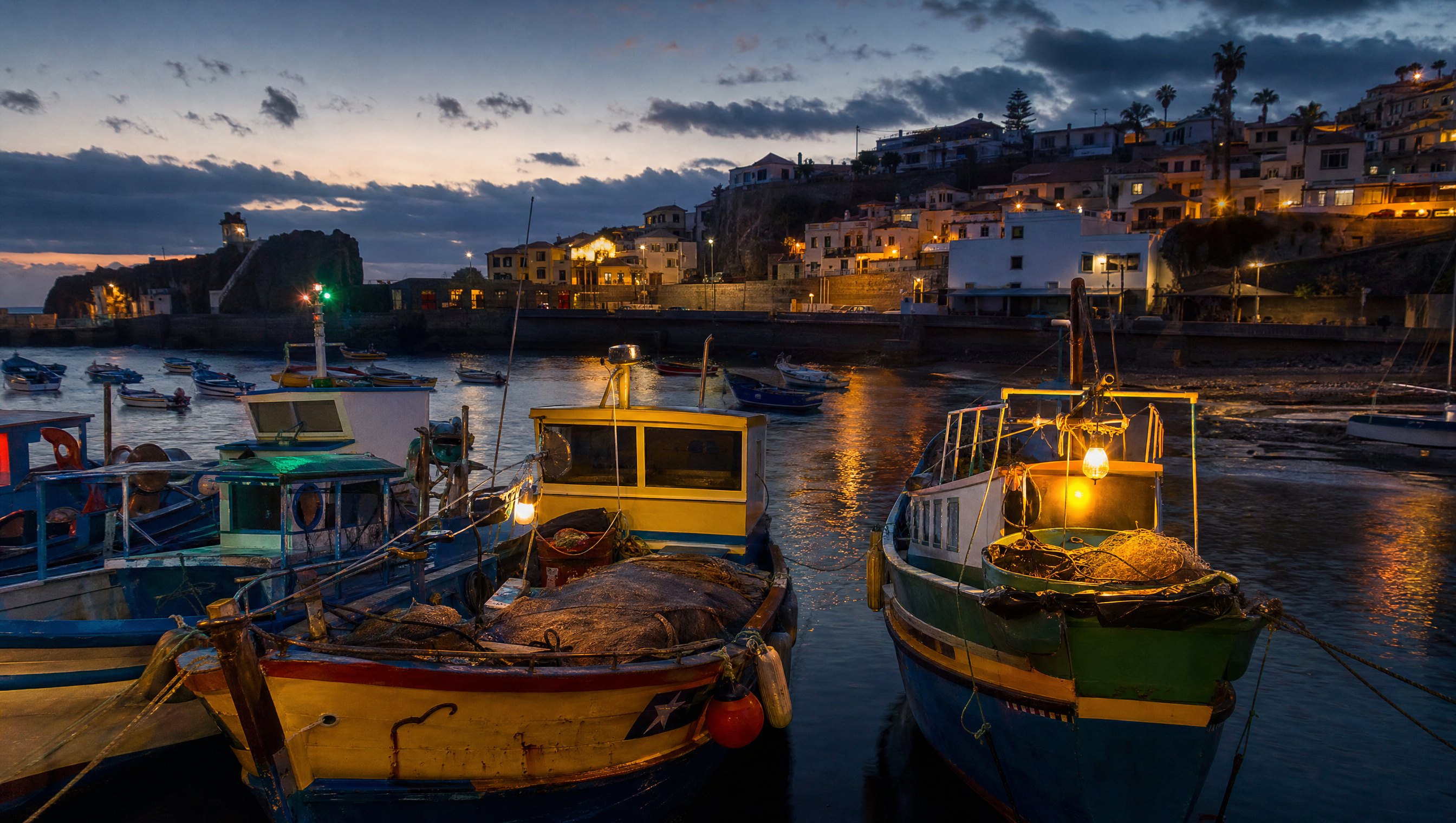 Des barques de pêche en bois colorées amarrées dans le petit port de Câmara de Lobos à la nuit tombante, lanternes allumées, filets et lignes de pêche, reflets sur l'eau sombre, ambiance de départ pour la pêche de nuit, photographie éditoriale réaliste