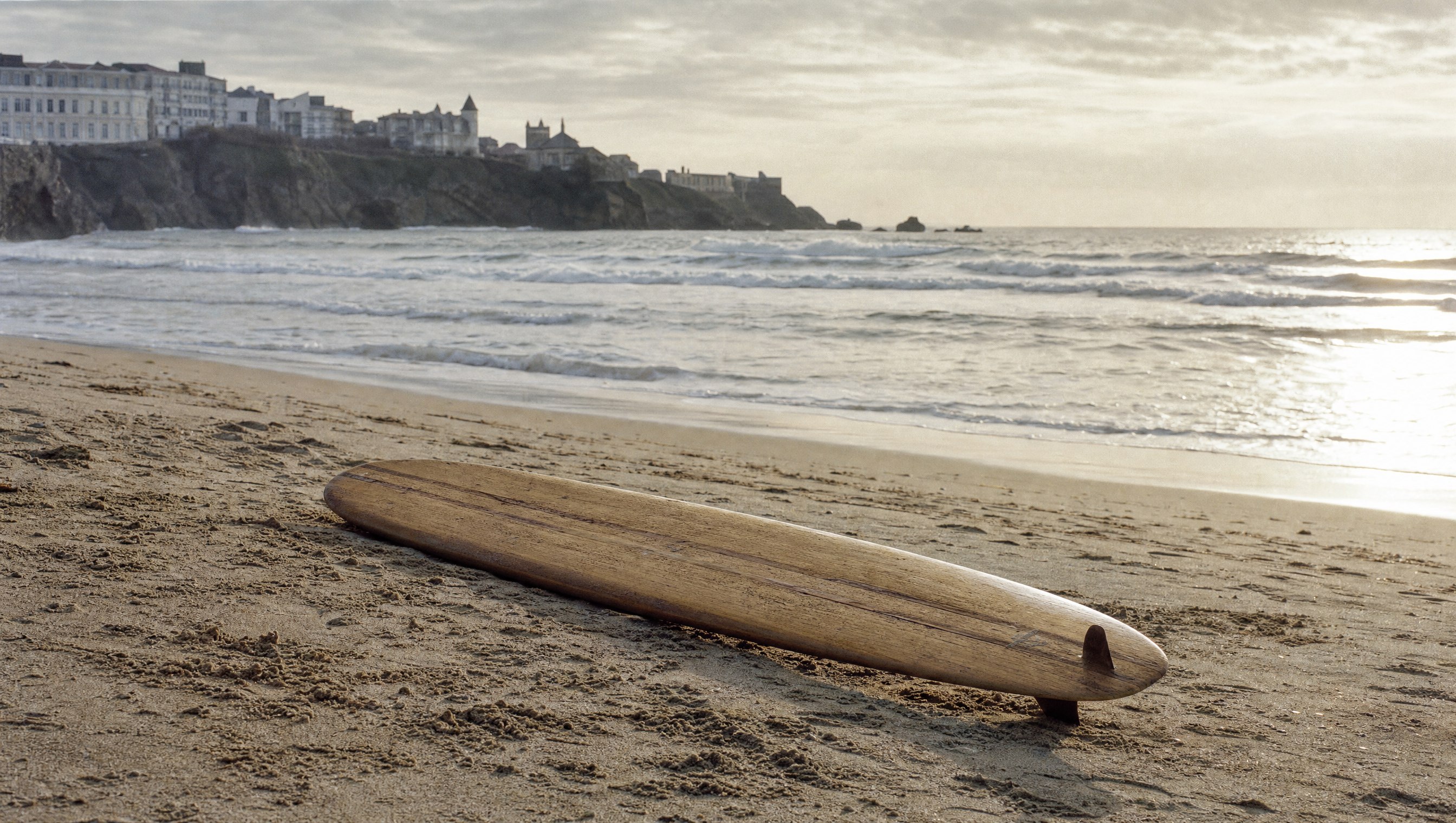 Une ancienne planche de surf en bois de balsa posée seule sur le sable de la Côte des Basques à Biarritz au petit matin, aucune personne, vagues et lumière argentée de l’Atlantique en arrière-plan, ambiance des années 1950
