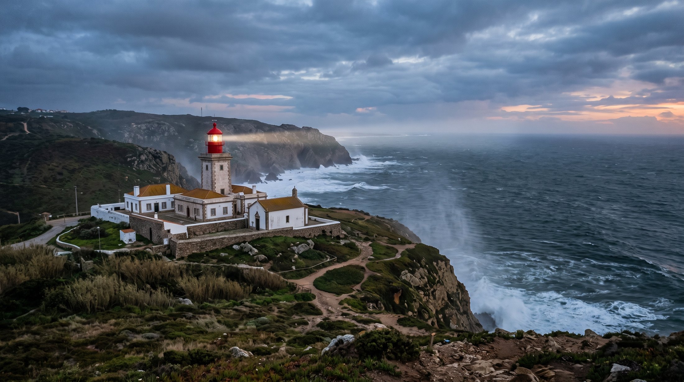 Phare du Cabo da Roca, tour trapue et bâtiments blancs à toiture ocre sur la lande, faisceau tourné vers l'océan au crépuscule, embruns dans l'air