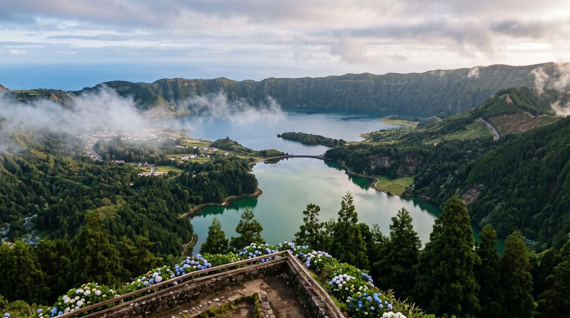 Vue plongeante sur la caldeira de Sete Cidades depuis le belvédère de Vista do Rei, brume se dissipant, deux lacs vert et bleu au fond du cratère, versants couverts de cryptomerias, lumière matinale douce