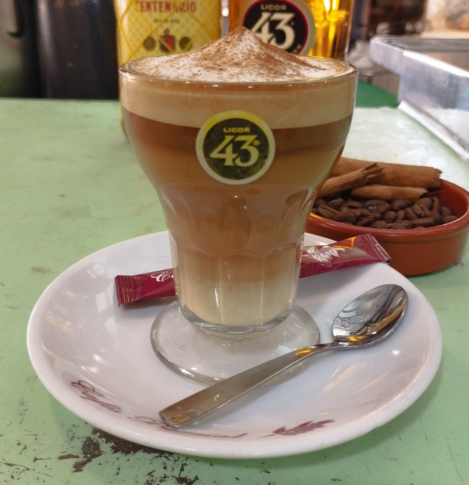Un café asiático de Cartagena dans son verre de cristal gravé, couches visibles de lait concentré, liqueur dorée et café, cannelle et zeste de citron sur le dessus, comptoir de bar en bois du quartier de pêcheurs de Santa Lucía, lumière chaude de midi