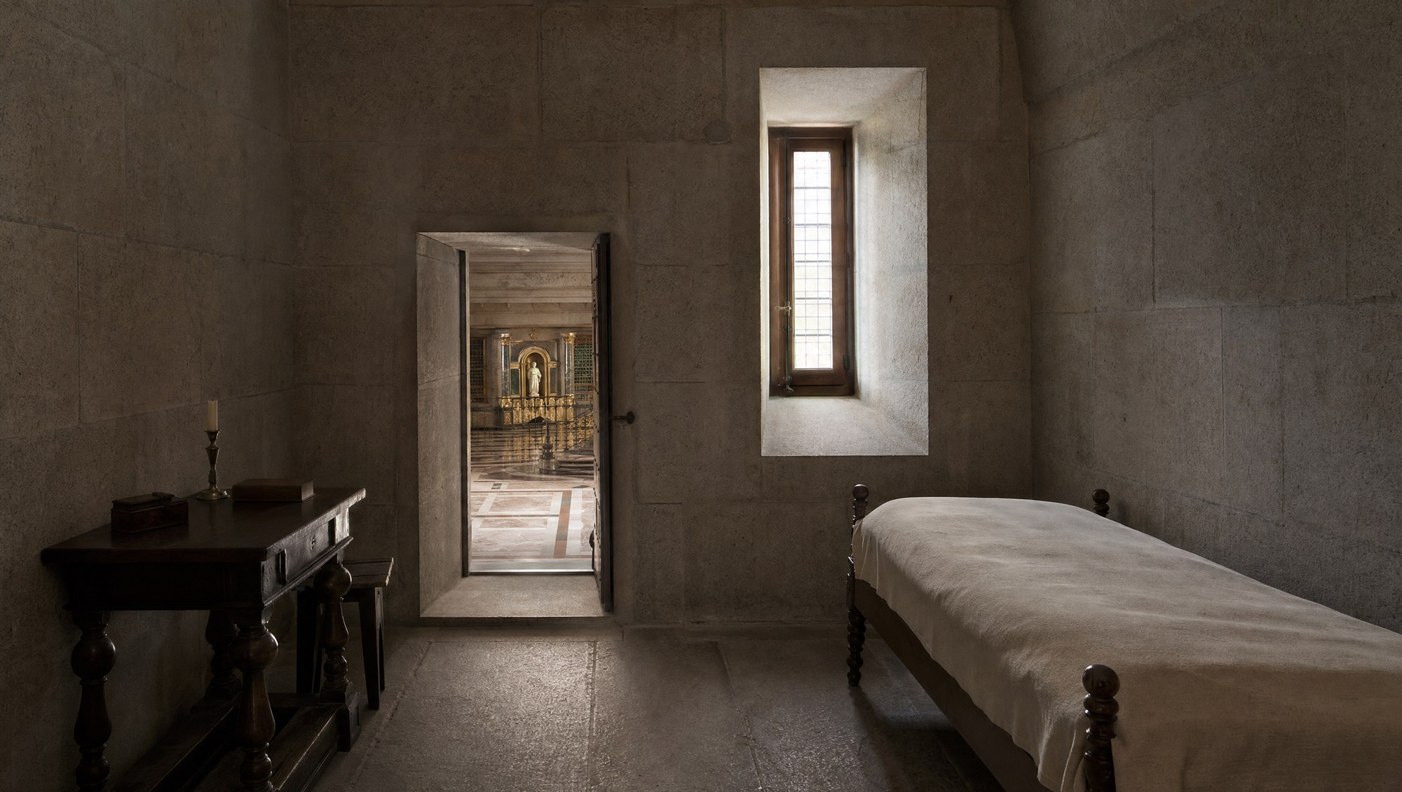 Chambre austère de Philippe II à l'Escorial, lit simple, petite table de travail en bois sombre et porte ouvrant sur la basilique, lumière tamisée entrant par une fenêtre étroite
