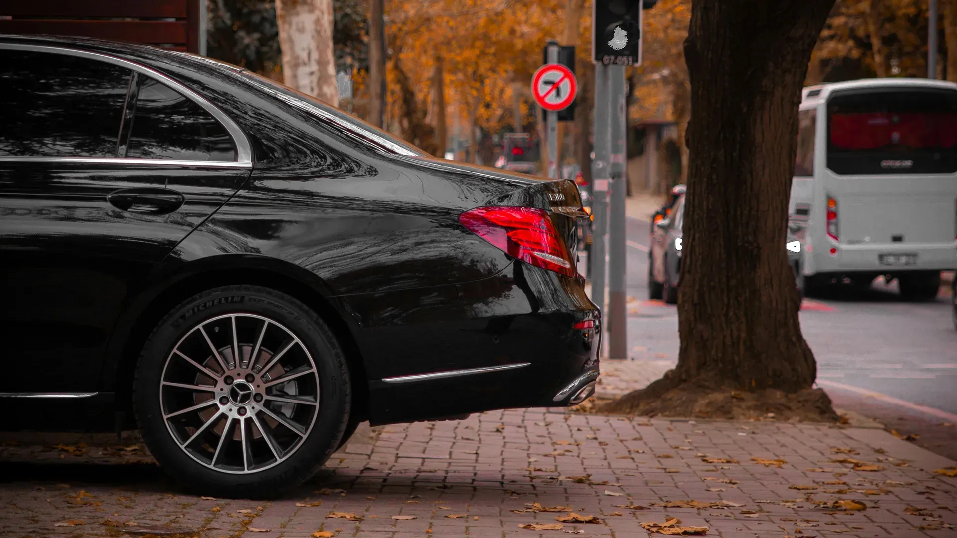 Rear side of a black Mercedes-Benz parked on a cobblestone street with autumn leaves scattered around and trees nearby.