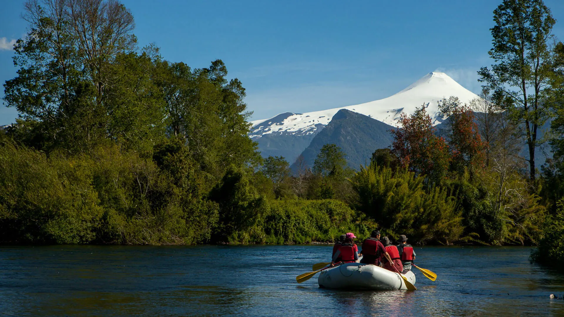 A raft with people floats down a river with a snow-covered volcano in the background