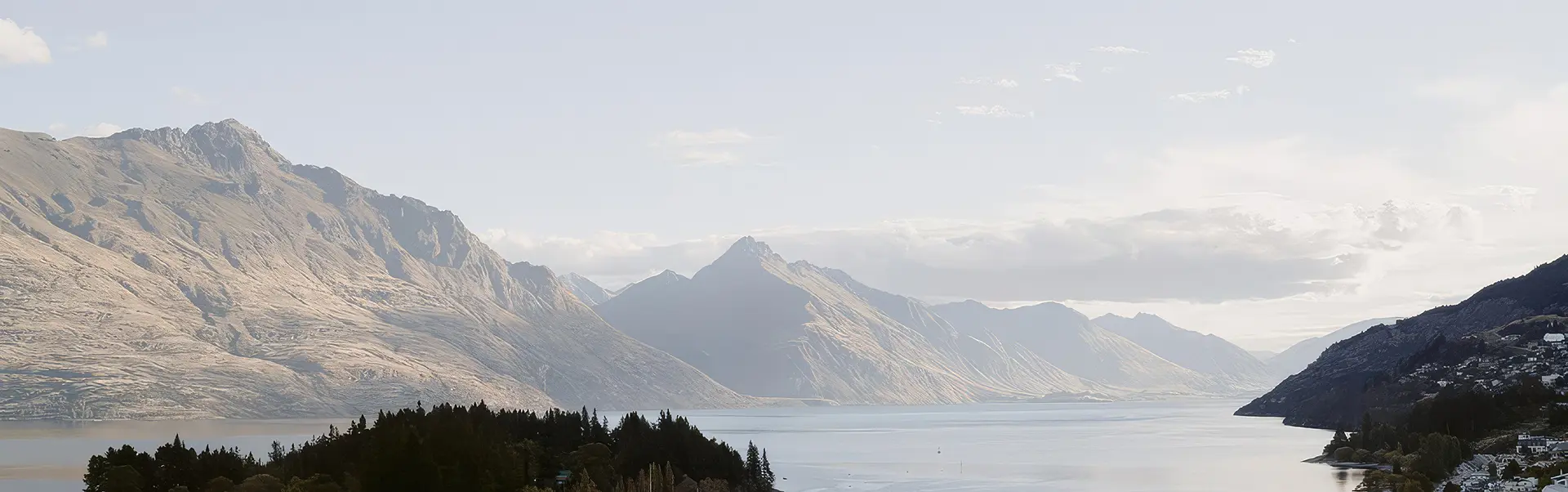 Wide lake surrounded by tall, rugged mountains under a pale, cloudy sky.