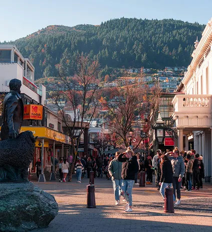Crowded pedestrian street in Queenstown with mountain and houses in the background under clear sky.