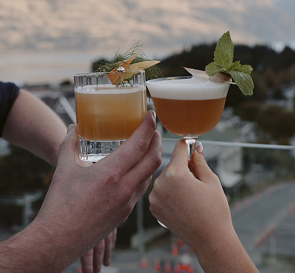 The Carlin - Two hands holding stylish cocktails garnished with herbs and citrus peel against a blurred outdoor background.