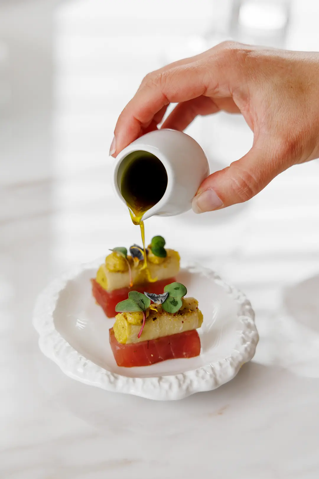 Hand pouring olive oil from a small white pitcher onto gourmet appetizers with garnish on a white plate.
