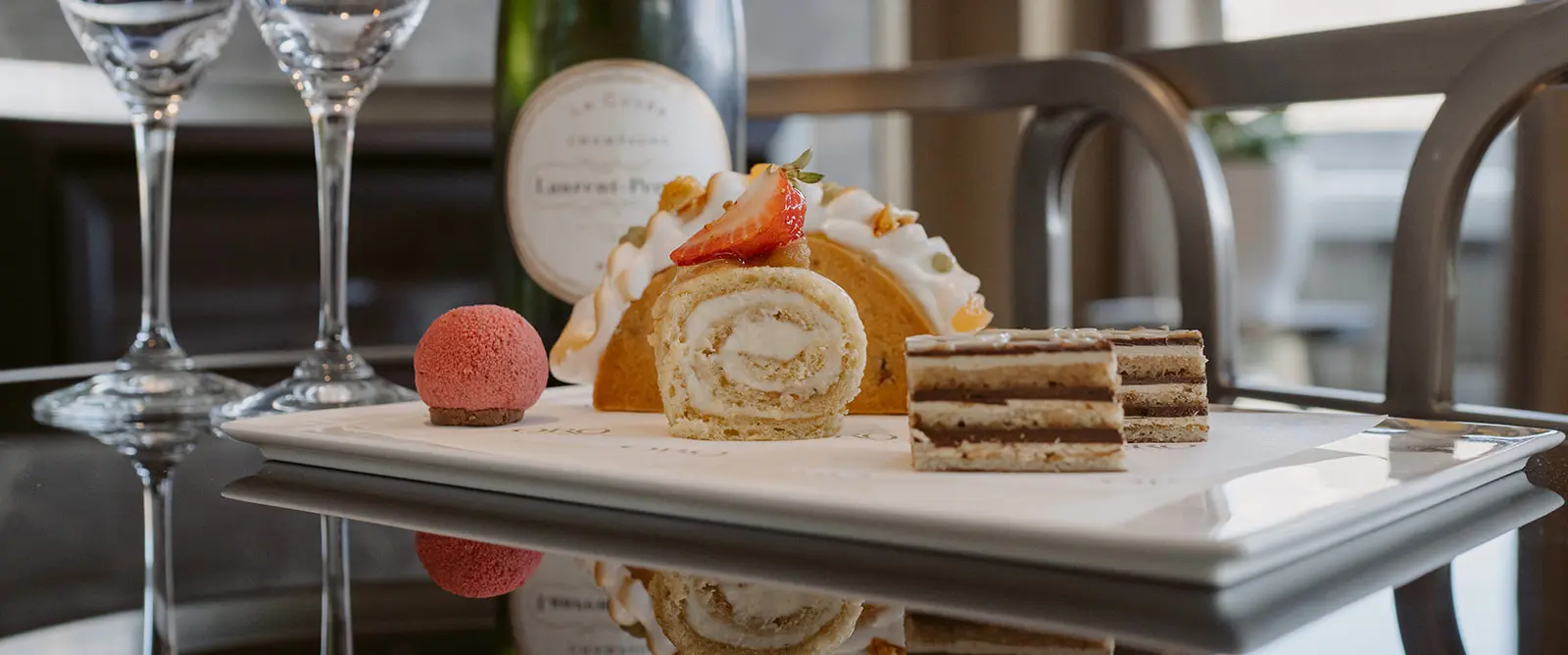 The Carlin - Plated assorted desserts including a strawberry-topped Swiss roll, a pink spherical dessert, layered chocolate cake slices, with a bottle of wine and empty wine glasses in the background.