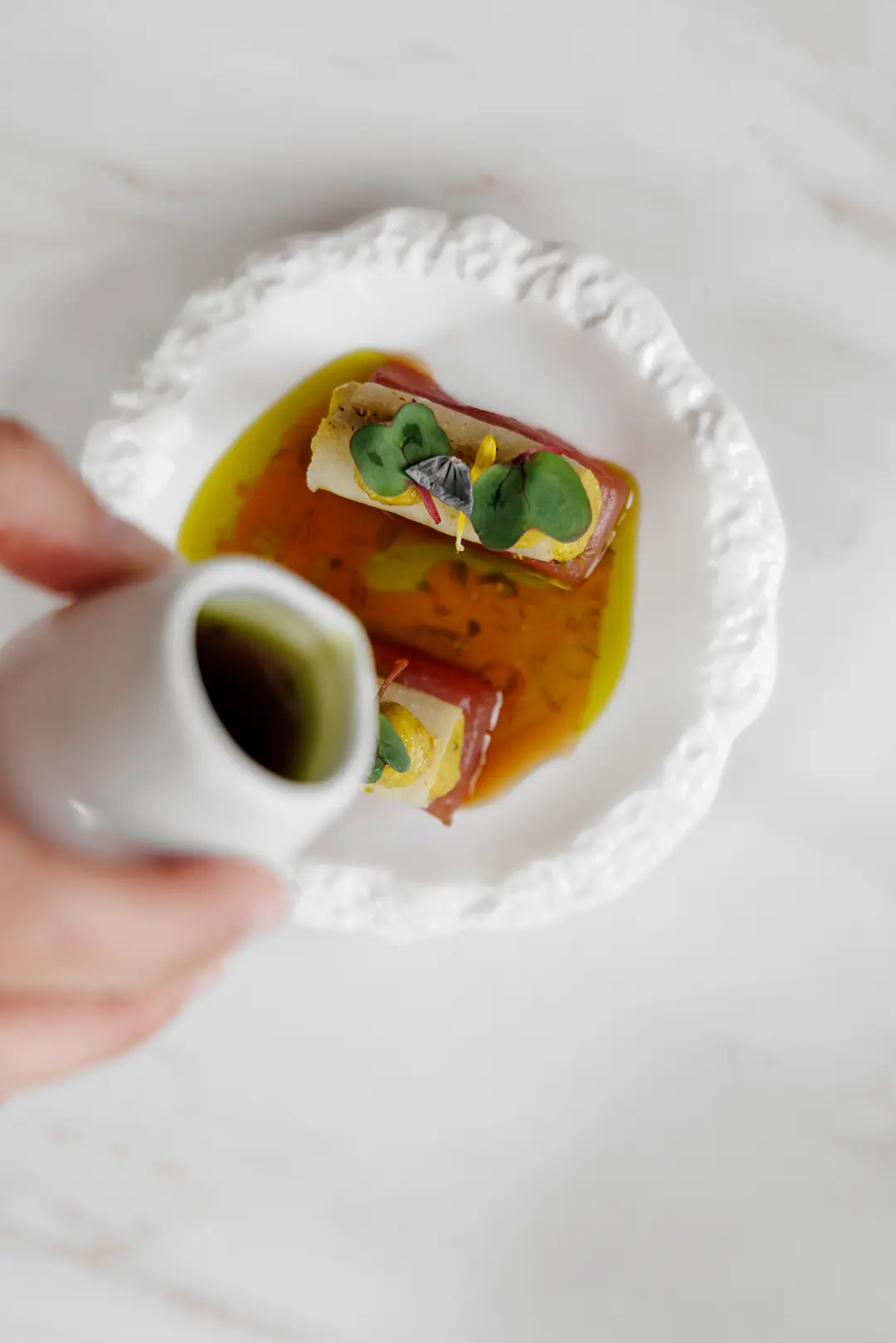 Two gourmet bites garnished with microgreens on a white ornate plate with a hand pouring green sauce onto the dish.