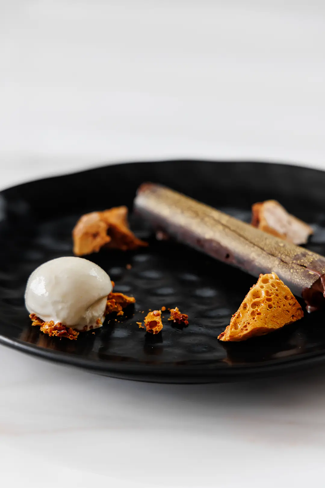 Artfully plated dessert with a scoop of ice cream, honeycomb pieces, and a chocolate stick on a black textured plate.
