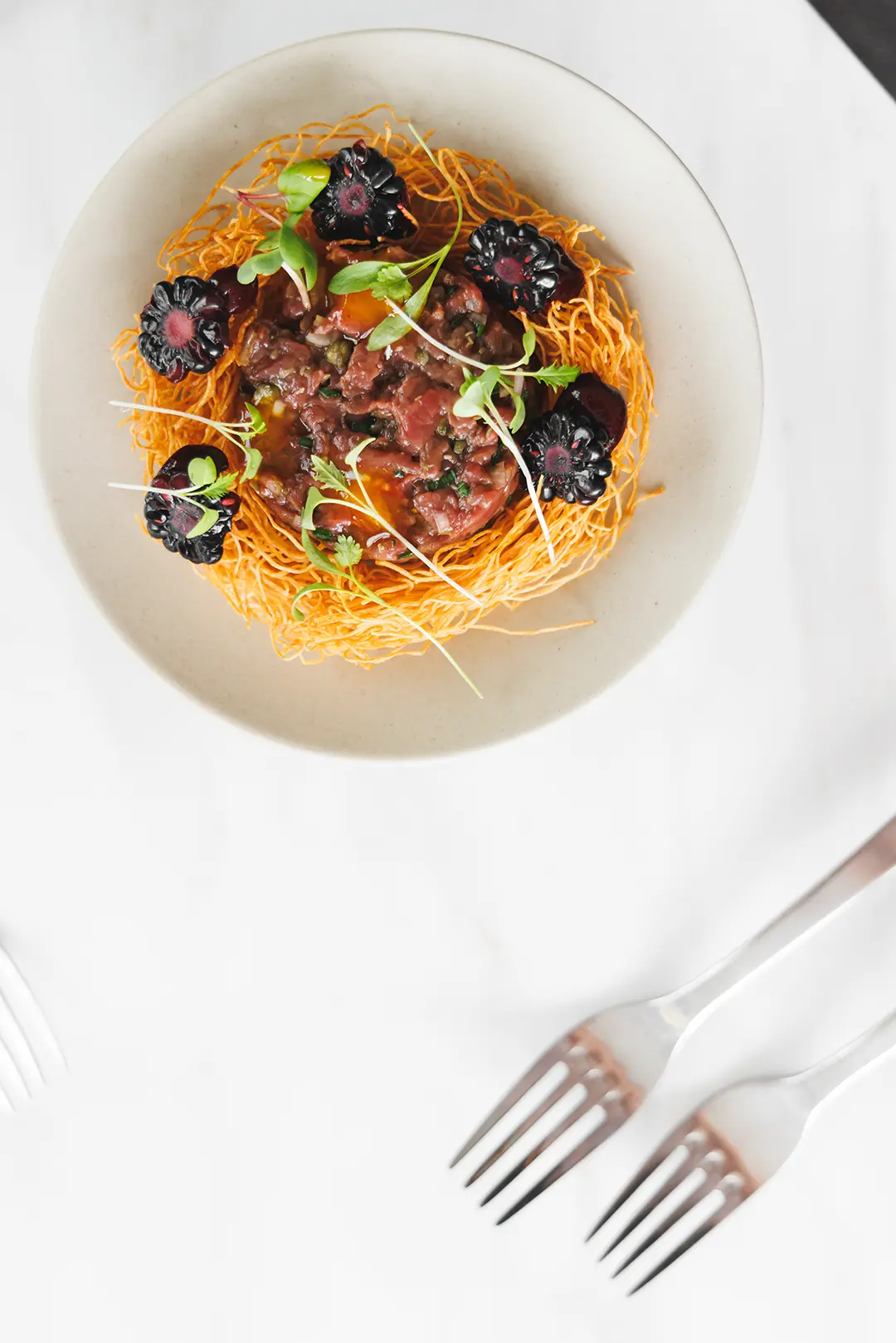 Gourmet dish with a nest of crispy orange noodles topped with diced meat, black corn, and microgreens on a beige plate.