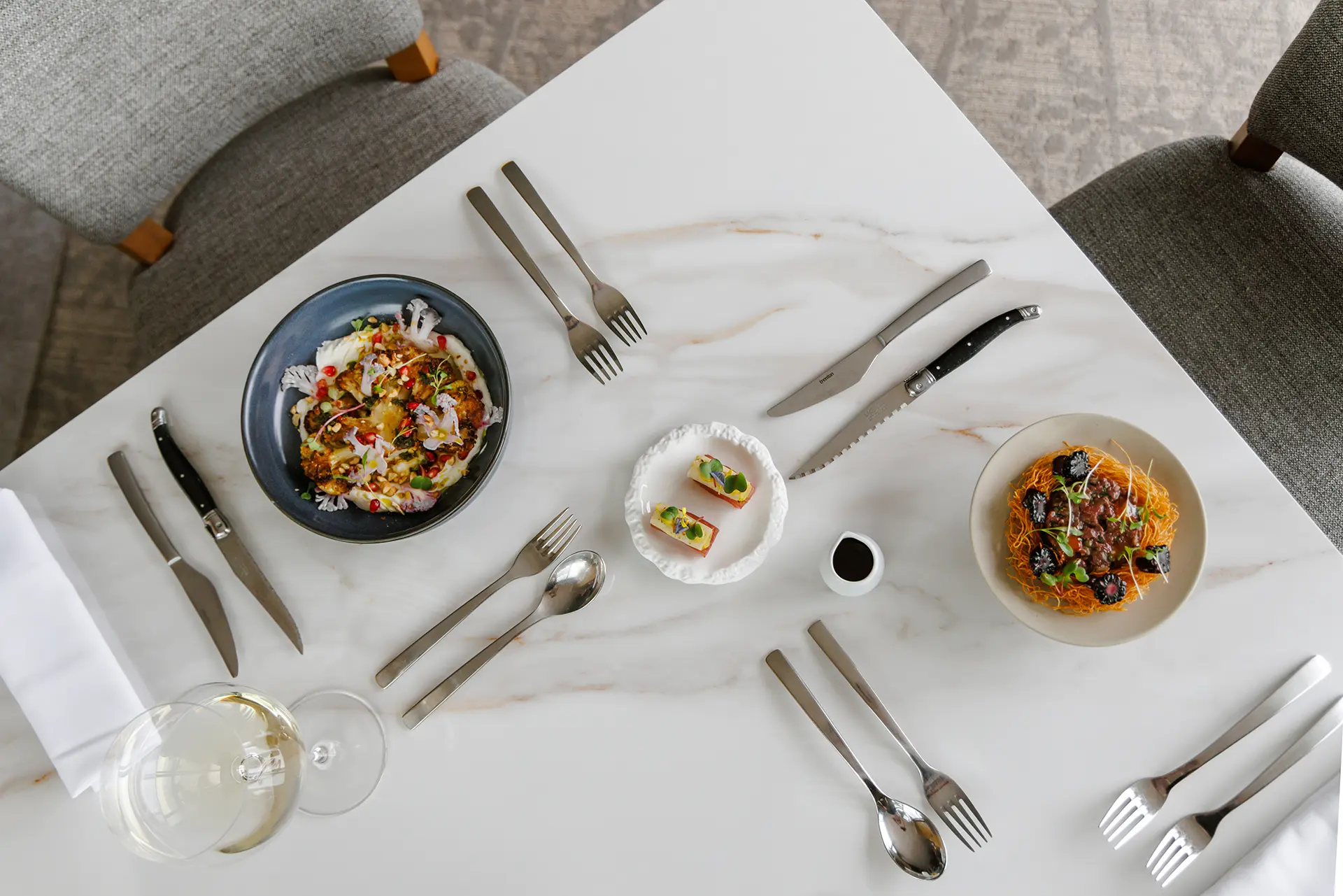 Overhead view of a white marble table set for two with gourmet dishes including a bowl of colorful cauliflower and pomegranate, a plate with small topped bites, and a dish of noodles with dark accents, alongside utensils and a glass of white wine.