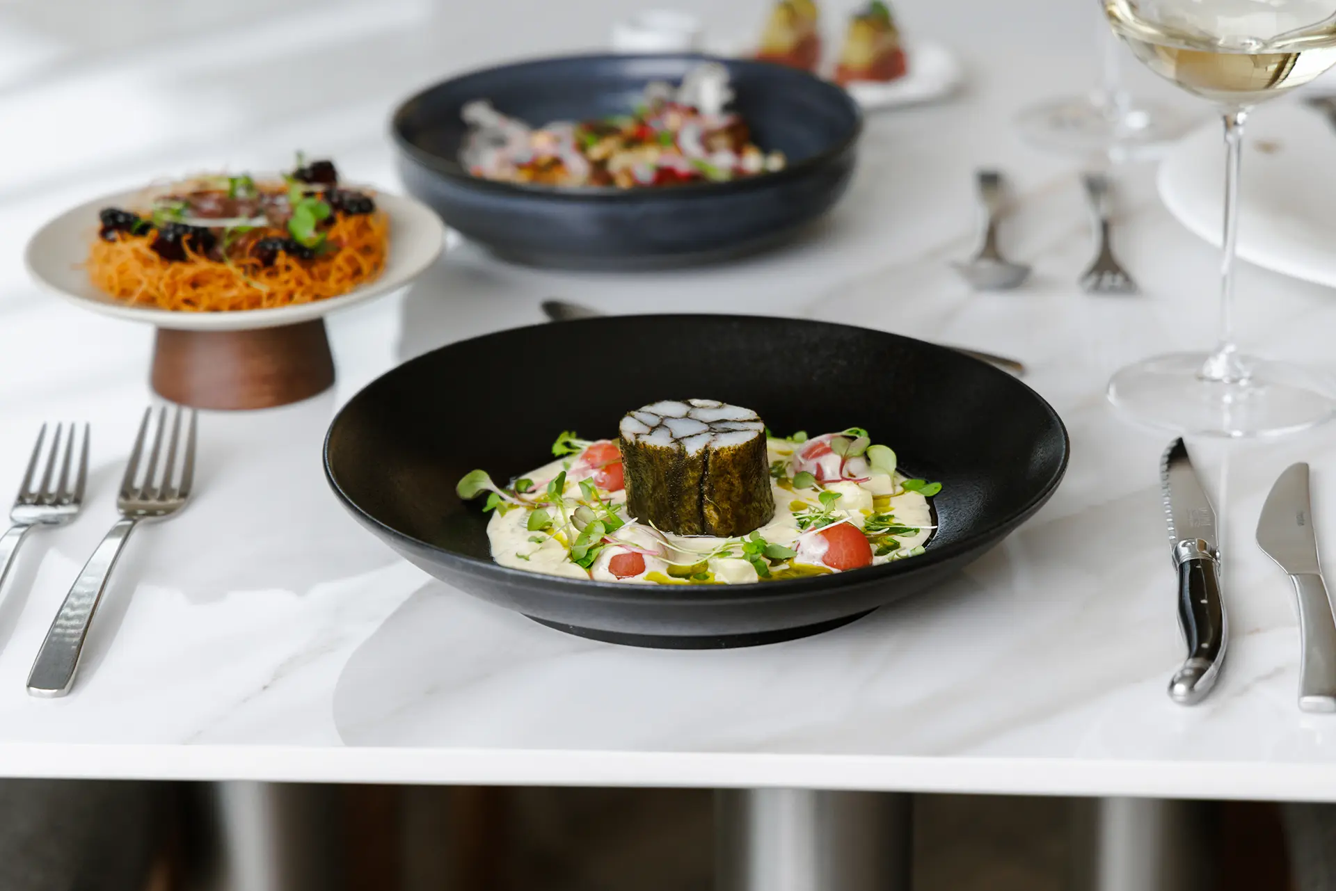 Gourmet dish with a cylindrical seaweed-wrapped fish centerpiece, surrounded by creamy sauce, cherry tomatoes, and microgreens on a black plate.