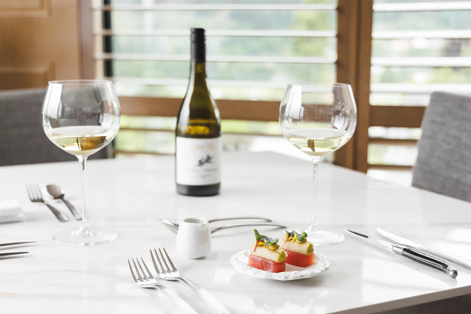 Elegant table setting with two glasses of white wine, a bottle of wine, and a small plate of gourmet appetizers on a white marble table.