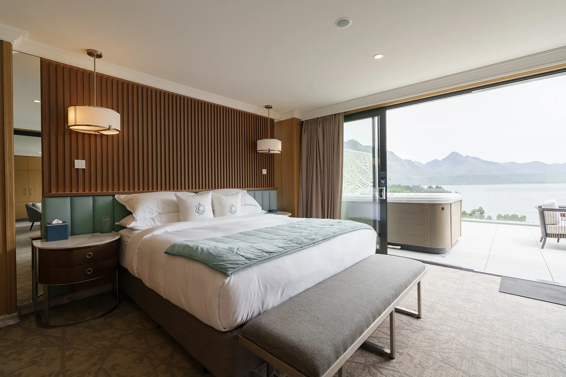 Modern hotel bedroom with a large bed, bedside tables, hanging lamps, bench, and sliding glass door opening to a balcony with a hot tub and mountain lake view.