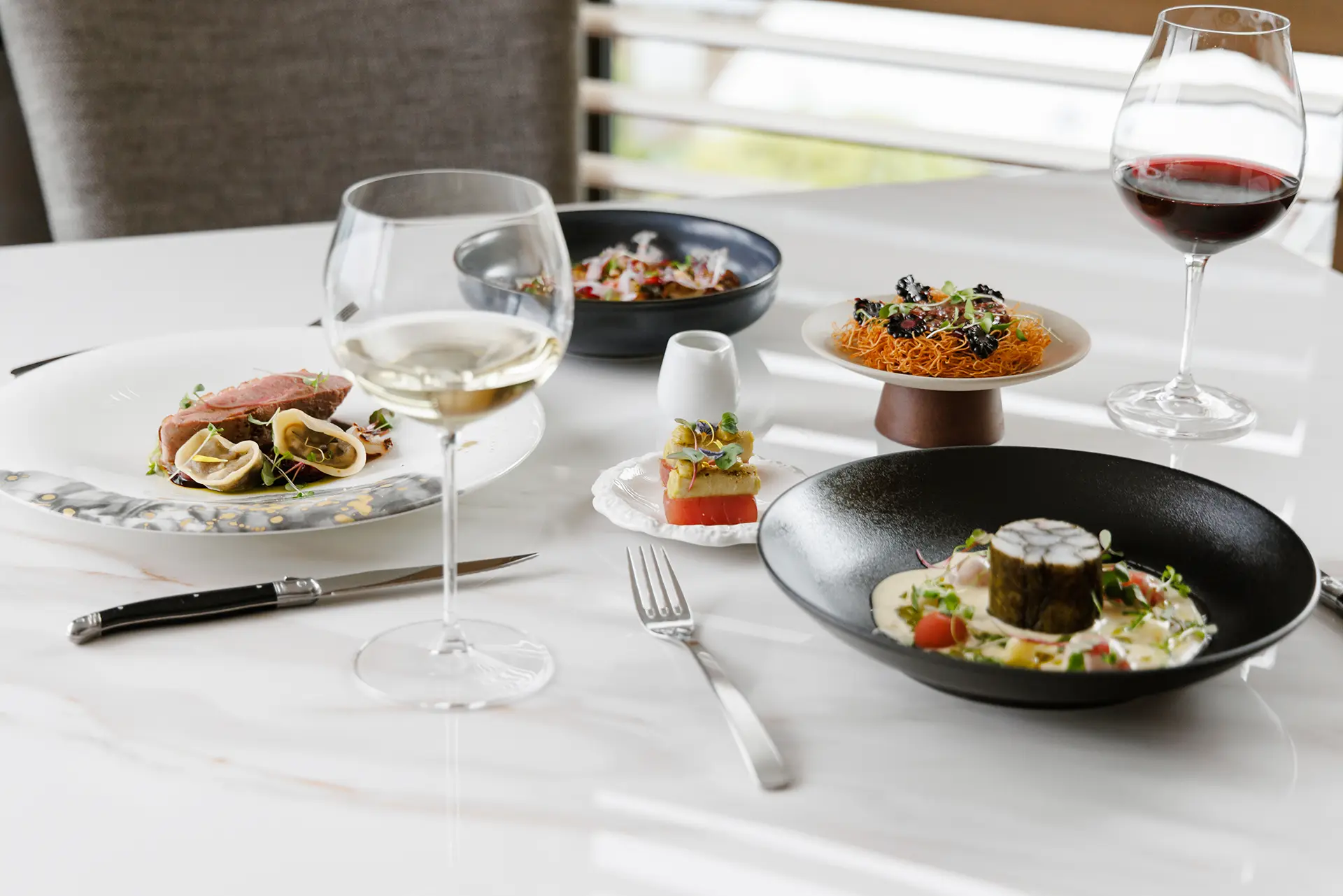 Gourmet dishes with two glasses of wine set on a white marble table in a fine dining setting.