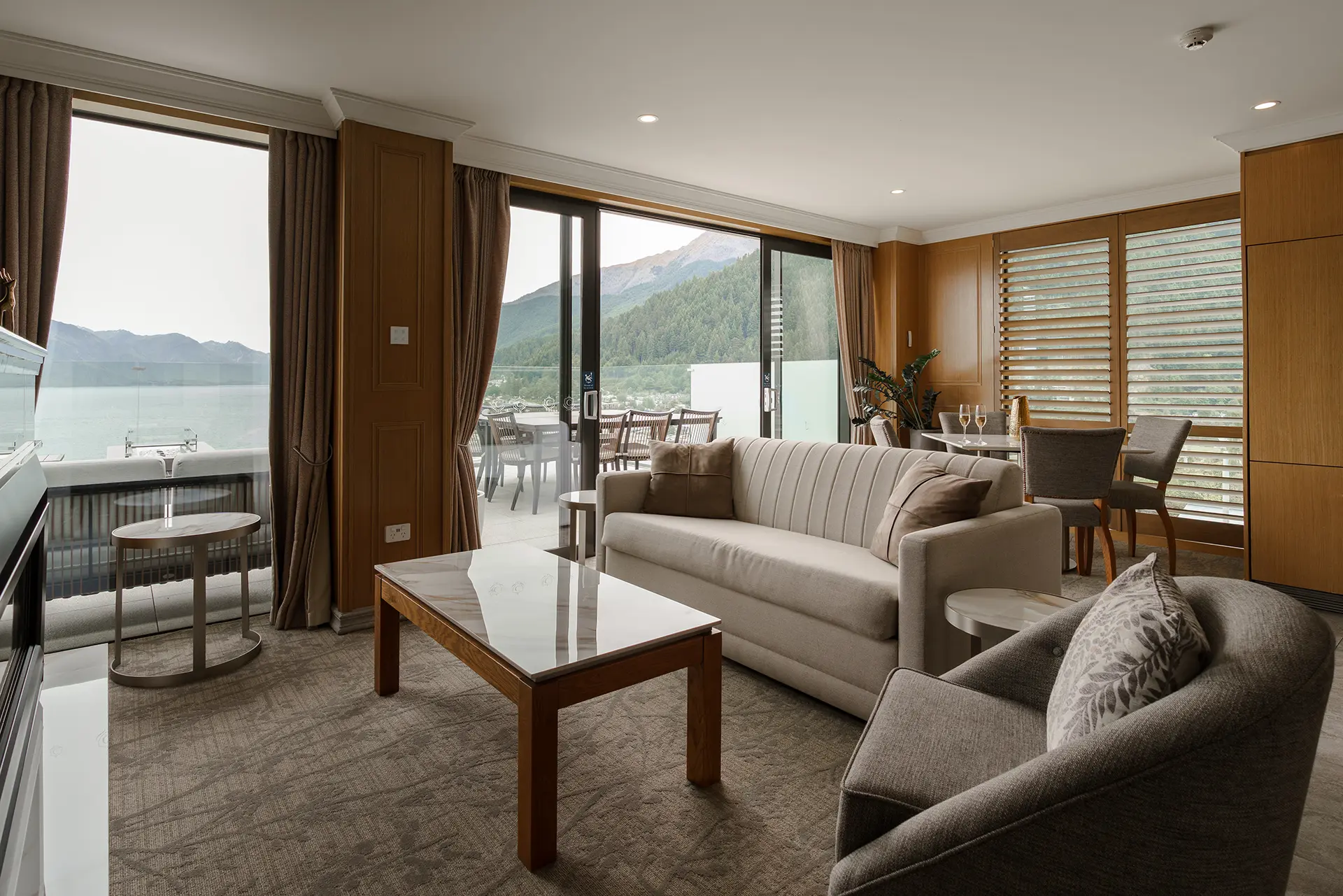 The Carlin - Modern living room with beige sofa, armchair, coffee table, and sliding glass doors opening to a balcony with mountain and lake view.