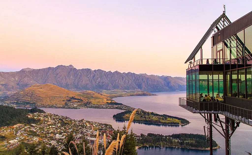 The Carlin - Scenic view of a lakeside town with mountains in the background and a glass-walled building overlooking the water during sunset.