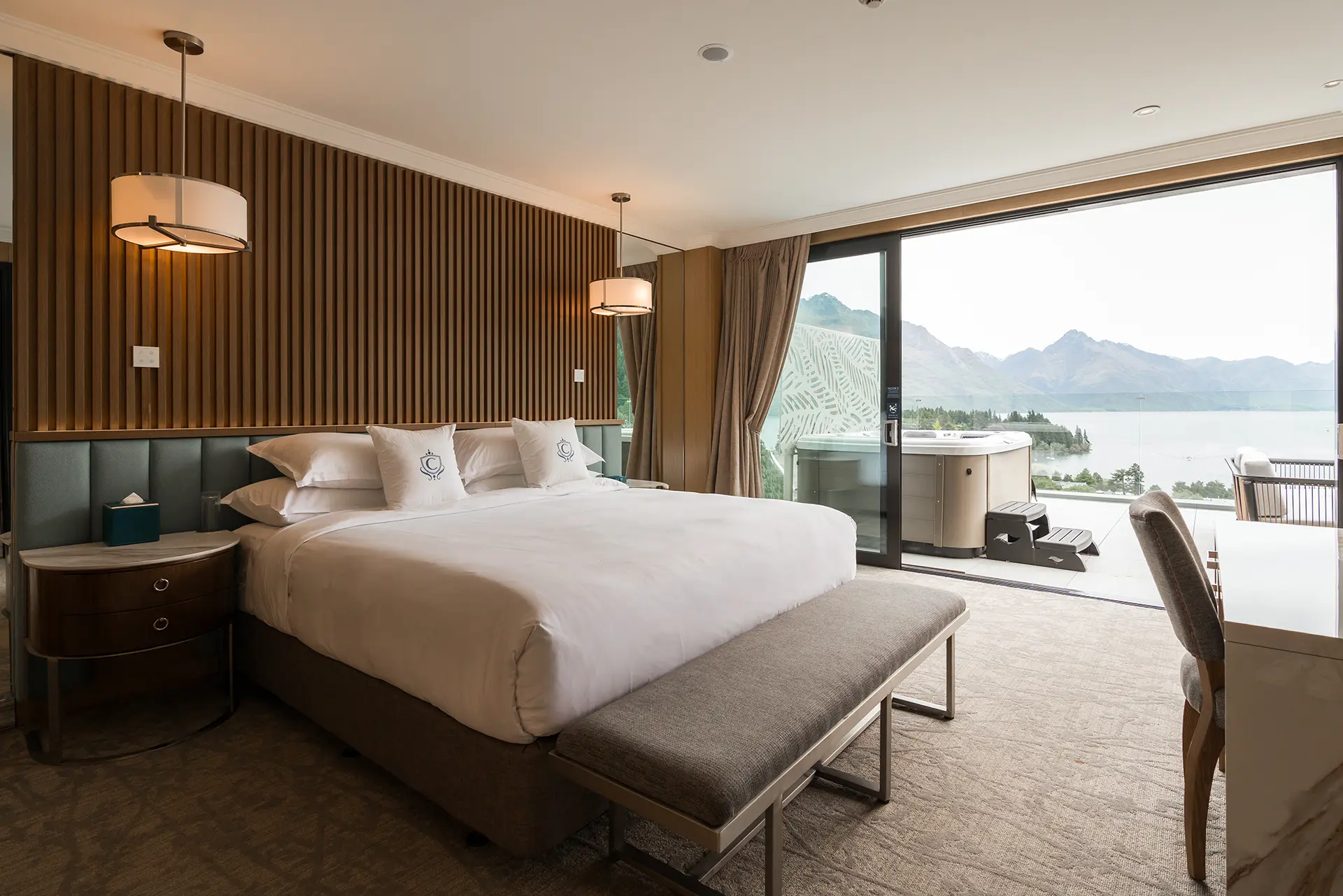 The Carlin - Modern hotel bedroom with large bed, the seating bench at foot, pendant lights, and sliding glass door opening to balcony with hot tub and mountain lake view.