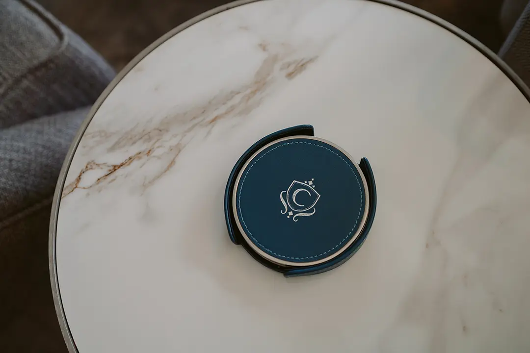 The Carlin - Round leather coaster holder with blue coasters featuring a white logo on a white marble table.