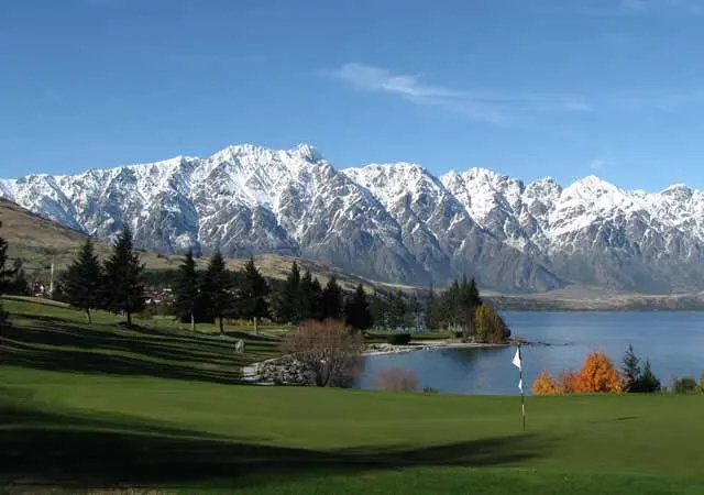 The Carlin Hotel - Golf course with a putting green and flagstick beside a lake, backed by snow-covered mountain peaks under a clear blue sky.