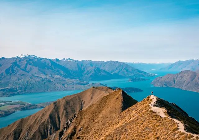 The Carlin Hotel - Hiker standing on a grassy mountain ridge overlooking a blue lake surrounded by mountain ranges under a clear sky.